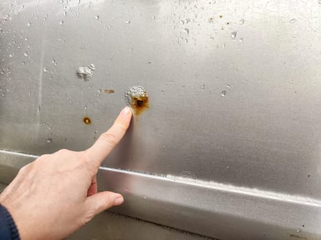 A person examines rust and scratches on silver car parked in a bright outdoor setting, focusing on the damaged area
