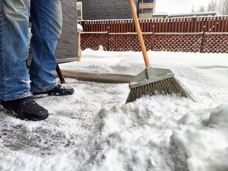 Individual uses a broom to clear snow from the driveway in a residential neighborhood during winter, showcasing heavy snowfall accumulation.
