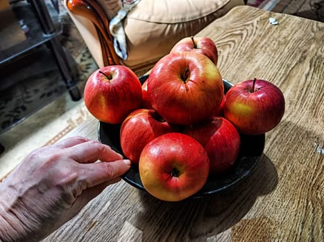A hand reaches for a bright red apple in a bowl filled with shiny, fresh apples on a rustic wooden table.