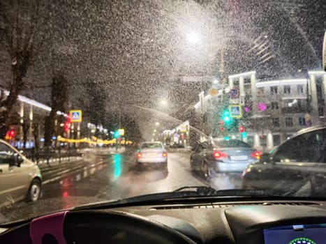 Drivers maneuver through a busy city street at night as colorful lights reflect on wet pavement, creating a lively urban scene.