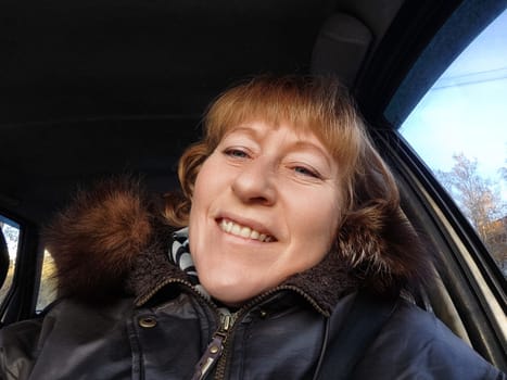 A middle-aged woman with blonde hair takes a cheerful selfie in her car while embarking on an autumn excursion and enjoying her journey.