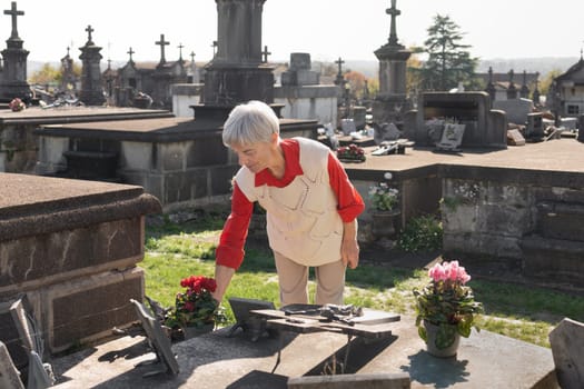 A grey-haired old woman lays flowers on her husband's grave in a Catholic cemetery