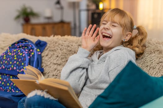 Young teenager girl taking a fairy tale book from backpack at home, flipping reading pages with interest. Blonde child kid on sofa pauses to admire illustrations, grinning at each charming scene.
