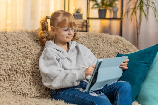 Young teenager girl draws colorful image at home, carefully moving stylus on tablet with very focused delight. Blonde child kid on sofa admires final creation, smiling proudly at bright digital art.