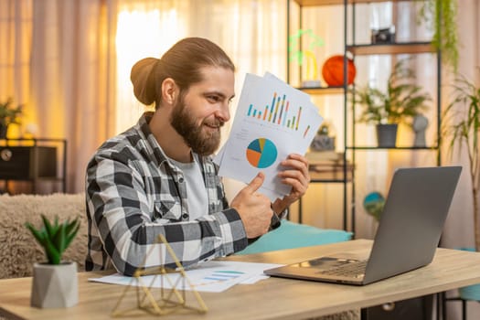 Young man starts laptop video chat at home table showing paperwork through webcam with active gestures. Caucasian guy on sofa smiling happily after hearing positive feedback or good result of work.