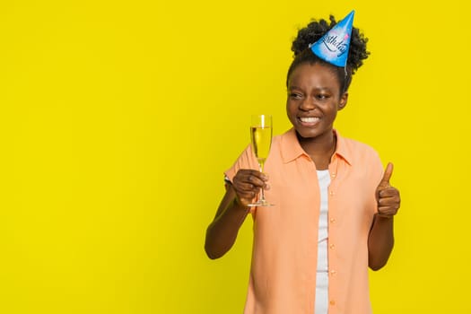 African American woman raises champagne glass while wearing festive party hat signaling a toast. Black girl isolated on yellow background emphasizes a celebratory moment for a special birthday event