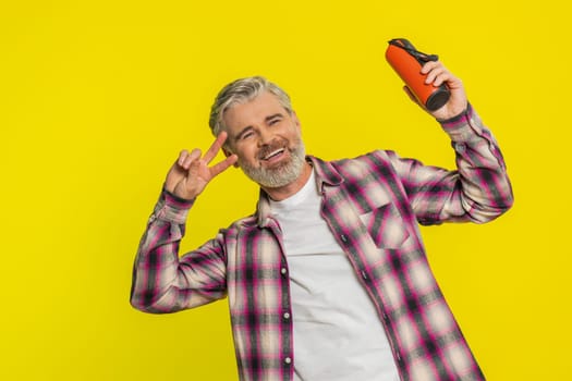 Mature man holds portable speaker listening music, dancing eagerly to favorite tunes, open delight. Middle-aged guy on yellow background radiates uplifting energy, emphasizing carefree music vibe.