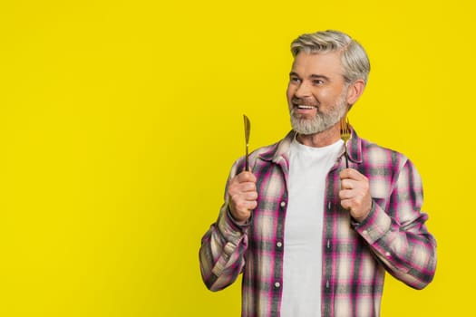 Ready to eat. Hungry Caucasian middle-aged man waiting for serving dinner dishes with with restlessness holding cutlery fork knife, will appreciate delicious restaurant meal on yellow background