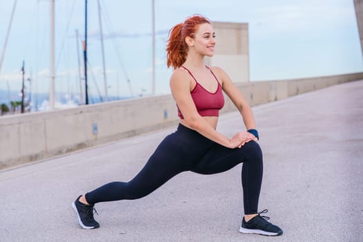 Athletic red-haired woman wearing sportswear performing a lunge stretch in an outdoor urban environment. She maintains balance and flexibility while preparing for exercise under a bright sky.