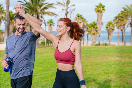 Smiling young man and woman celebrating after exercising outdoors. They are wearing sportswear and enjoying a joyful moment together in a green park with palm trees near the sea.