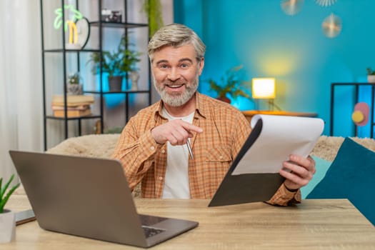 Caucasian mature man sitting at home table reviewing important documents and using laptop. Middle-aged guy smiles with satisfaction from successful work, demonstrating productivity and accomplishment