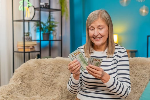 Happy Caucasian mature woman counts money dollar cash in hands after winning lottery bonus or receiving a gift. Grandmother smiles broadly sitting on home sofa, celebrating financial victory salary