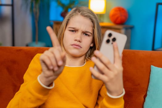 Teenager boy sits on sofa at home, waving hand in disagreement while looking unhappy at smartphone. Dissatisfied with game or app on phone, shows frustration disapproval, displaying negative emotions