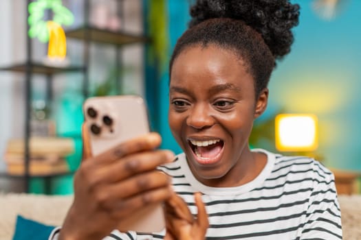 African American woman showing surprise after reading unexpected good happy message on smartphone. Sitting indoors on sofa, wide eyes, open mouth shocked expression, emotional reaction, focused moment