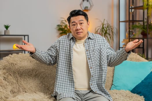 I dont know what to say. Confused Asian handsome man feeling embarrassed about ambiguous question, having doubts, no idea, being clueless and uncertain at home. Chinese guy sits on sofa in living room