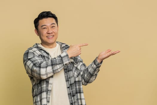 Asian middle-aged man showing thumbs up and pointing right empty place, advertising area for commercial discount text, copy space for goods promotion advertisement. Chinese guy on beige background