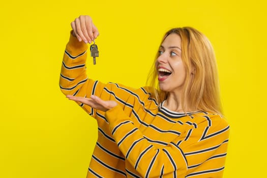 Caucasian young blonde woman real estate agent lifting hand showing the keys of new home house apartment, buying or renting property, mortgage loan, insurance. Girl isolated on yellow background