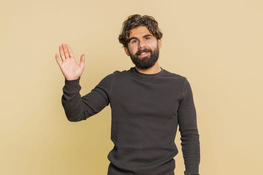 Hello, welcome. Handsome young Indian man with long sleeves waving hi with his palm, greeting with hospitable friendly smile. Adult male Hispanic guy isolated on beige studio background indoors.