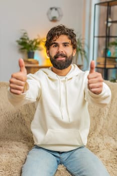 Like. Happy excited Indian young man looking at camera and showing thumbs up sign positive great news positive feedback. Bearded Hindu guy in hoodie sitting on sofa couch in living room at home.