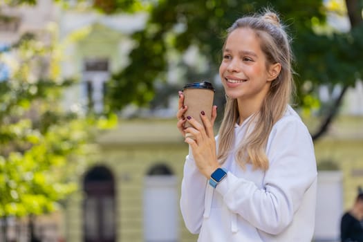 Happy smiling Caucasian woman enjoying morning coffee hot drink. Relaxing, taking a break. Young girl teenager walking through urban city center street, drinking coffee to go. Town lifestyles outside
