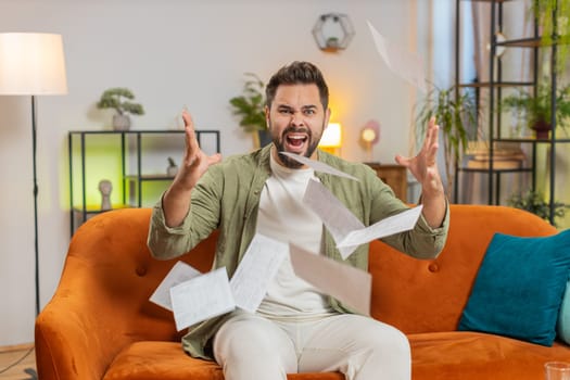 Stressed brunette man in living room looking at unpaid bank debt bills, doing paperwork, planning budget, calculate finances mortgage payments, deadline. Guy throws paper bills at home apartment.