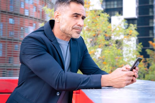 A middle-aged businessman in a stylish suit leaning on a railing, holding his smartphone with a focused expression. The modern city background reflects a professional and contemplative mood.