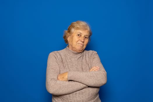 Caucasian woman about 70s wearing dark beige sweater shrugging shoulders, feeling confused and unsure, doubting with arms crossed. Isolated over blue background.