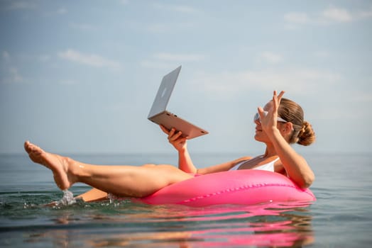 A woman is floating on a pink inflatable raft in the ocean while holding a laptop. Concept of relaxation and leisure, as the woman is enjoying her time in the water while working on her laptop