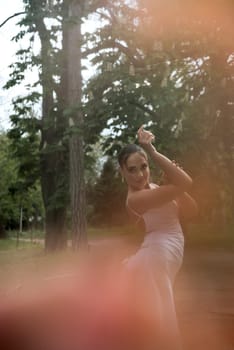 elegant girl in a long white dress against the background of trees. white dress fits her figure, brunette with a smooth hairstyle and tanned skin, rainbow in the frame, pink haze