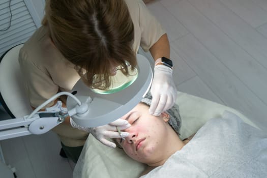 A cosmetologist examines a patient's face using a magnifying lamp during a skin treatment. The patient lies supine on a treatment table.