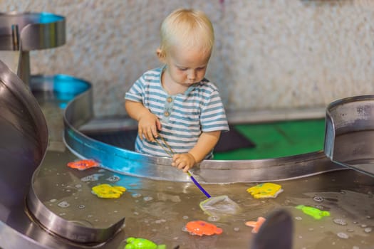 Toddler boy catching toy fish with a net in a vibrant water play area at an entertainment center. A joyful moment of learning, play, and discovery designed to inspire young children.