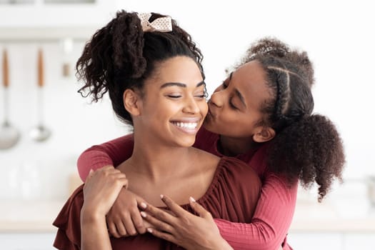 Cute black girl teenager hugging and kissing her happy beautiful mom after breakfast together, kitchen interior. Loving afro-american mother and daughter bonding while having snack, copy space