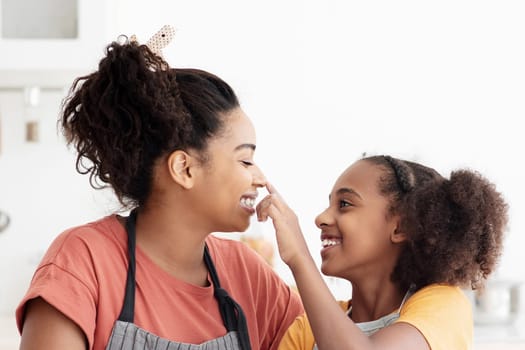 Happy afro-american mother and teen daughter in aprons having fun while baking, cheerful black mom and kid playing with flour while making dough for cookies at kitchen, enjoying time together