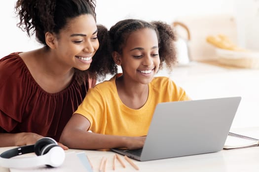 Cute black mother and kid using laptop together, sitting at kitchen table, hugging and looking at computer screen, spending time together, watching cartoon, copy space, closeup photo