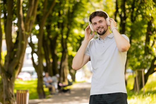 Happy young Caucasian man put on wireless modern headphones earphones. Runner jogger guy sport male run runner in park outdoors enjoy music listening song audio jogging healthy lifestyle fitness.