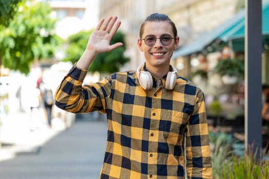 Cheerful young man tourist smiling friendly at camera, waving hands gesturing invitation hello, hi, greeting, goodbye, welcoming with hospitable expression outdoors. Guy on urban city town street.