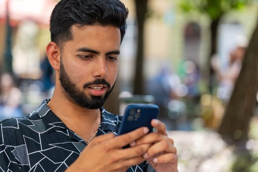 Indian young man using smartphone typing text messages in social media application online, surfing internet, relaxing, taking a break outdoors. Hispanic guy tourist sitting on chair in city street.