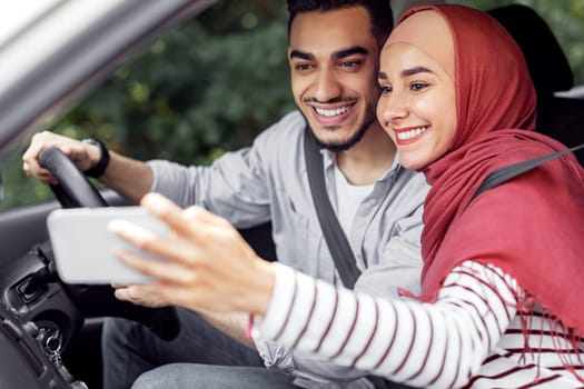 Happy young muslim female in hijab and husband driving with steering wheel, take break, make selfie or video call on phone in car. Mobile app, travel together and trip, auto vacation and online map