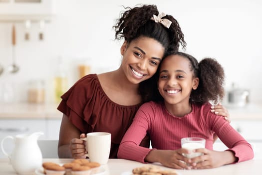 Loving young black mother and teen daughter enjoying fresh homemade cookies, drinking coffee and milk, sitting at tablet in kitchen and smiling at camera, spending time together at home, copy space
