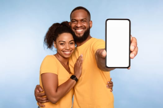 New awesome mobile app. African American couple presenting smartphone with mockup, promoting application or website, advertising your product or service, blue studio background. Selective focus
