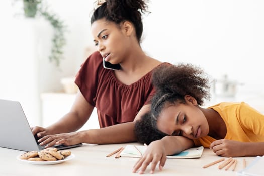 Bored african american teen girl sitting by her working mom, busy black lady having phone conversation and typing on laptop, upset daughter laying on table, looking for mother attention, copy space