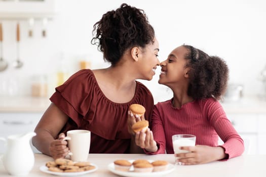 Loving african american mom and daughter enjoying time together at home, having snack, drinking milk, eating homemade pastry and bonding, touching each other with noses and smiling, copy space