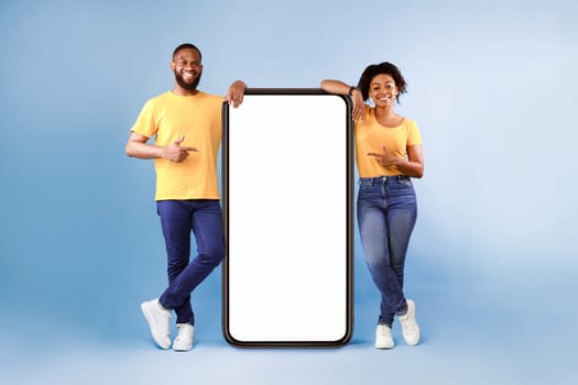 Great mobile app. African American couple pointing at giant smartphone with mockup, promoting application or website, advertising product or service, blue background. Full length