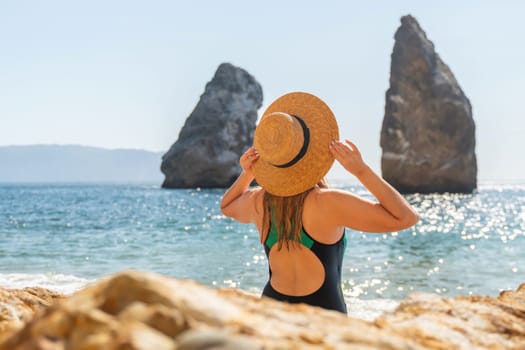 A woman in a swimsuit and hat stands on a beach, facing two large rock formations in the ocean.