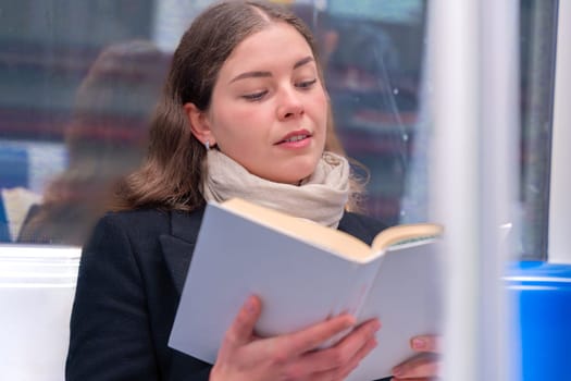 Young woman in a black coat and beige scarf sitting inside a modern subway train, reading a book with focus. She enjoys a quiet moment during her commute in the city.