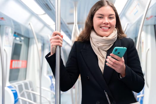 Young woman in a black coat and beige scarf standing inside a modern subway train, holding a smartphone and gripping a metal pole. She appears focused while commuting, carrying a stylish white purse.