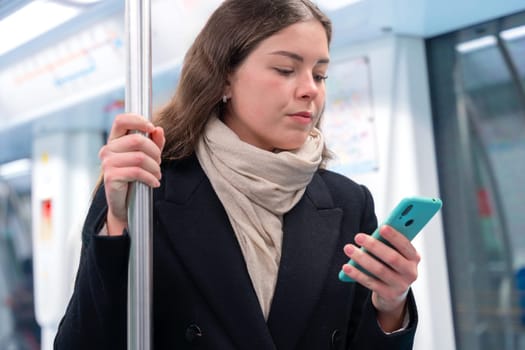 Young woman in a black coat and beige scarf standing inside a modern subway train, holding a smartphone and gripping a metal pole. She appears focused while commuting, carrying a stylish white purse.