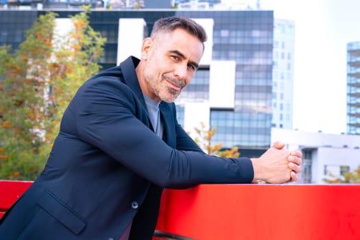 A middle-aged businessman in a stylish suit leaning on a railing and smiling. The modern city background enhances his confident, approachable, and professional demeanor.