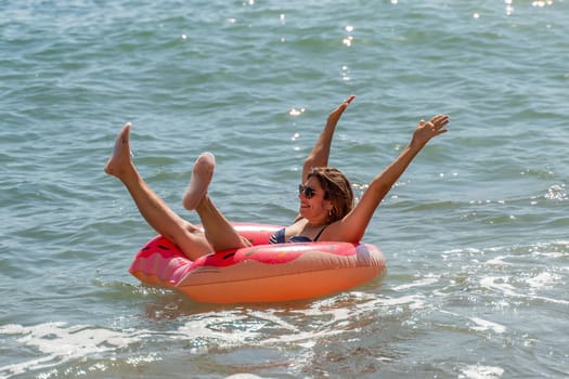 A woman in a pink inflatable ring floats on the ocean, arms raised in the air, enjoying a sunny day.