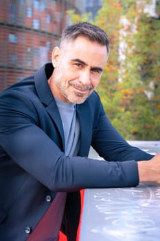 A middle-aged businessman in a stylish suit leaning on a railing and smiling. The modern city background enhances his confident, approachable, and professional demeanor.